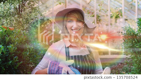 Watering shrubs gardener wearing sun hat, striped apron in greenhouse, holding blue watering can Watering shrubs gardener wearing sun hat, striped apron in greenhouse, holding blue watering can 135708524