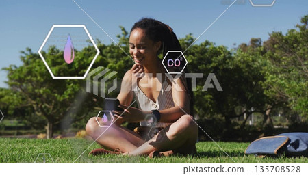 Smiling woman holding smartphone and wearing smartwatch while sitting on park lawn, with backpack 135708528