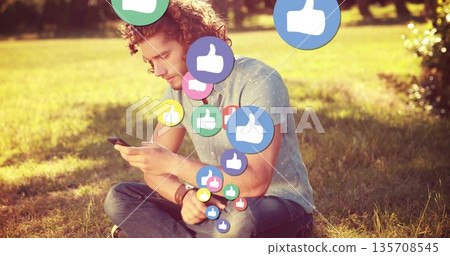 Sitting man wearing dotted shirt scrolling smartphone in park, with floating thumbs up icons 135708545