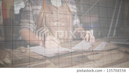 Sketching craftsman wearing leather apron drawing plans on paper at studio bench, with grid overlay Sketching craftsman wearing leather apron drawing plans on paper at studio bench, with grid overlay 135708556