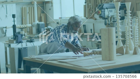 Leaning senior woodworker inspecting paper plans on workshop bench, with prototypes and drill press Leaning senior woodworker inspecting paper plans on workshop bench, with prototypes and drill press 135708566