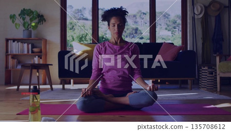 Adult woman in athletic wear practicing meditation on yoga mat in living room, with water bottle Adult woman in athletic wear practicing meditation on yoga mat in living room, with water bottle 135708612
