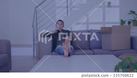 Reclining jeans-clad woman sipping dark bottle on sofa at home, with coffee table and moving box 135708622