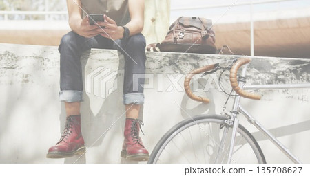 Sitting man in sleeveless top scrolling phone on concrete ledge in plaza with backpack bicycle 135708627