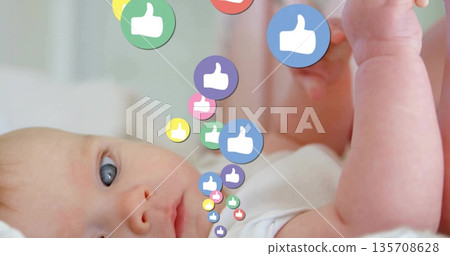 Infant wearing white onesie lying on bed in nursery, looking at colorful thumbs-up icons descending 135708628