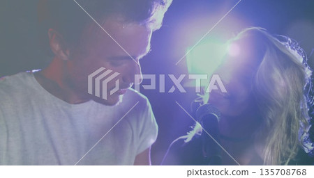 Singing duet partners wearing white dark outfits leaning toward mic on club stage, with lights haze 135708768