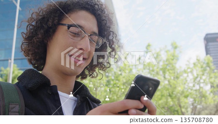 Checking smartphone, male student standing by glass facade on sidewalk, with backpack and lamppost Checking smartphone, male student standing by glass facade on sidewalk, with backpack and lamppost 135708780