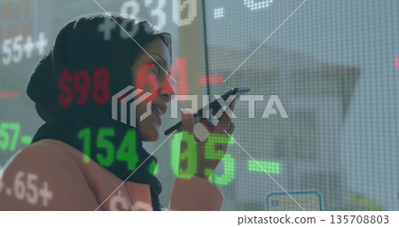 Executive wearing black hijab blazer speaking into smartphone in office by glass with data overlay Executive wearing black hijab blazer speaking into smartphone in office by glass with data overlay 135708803