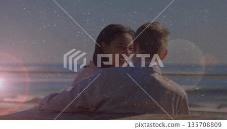 Laughing couple wearing pink and gray shirts hugging on bench at beach, with ocean, lens flare 135708809