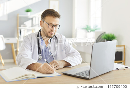 Focused serious young male doctor in white uniform working online on computer, improving Focused serious young male doctor in white uniform working online on computer, improving 135709400