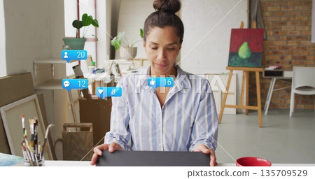 Holding tablet, mature woman viewing social icons in bright art studio, with brushes and canvas Holding tablet, mature woman viewing social icons in bright art studio, with brushes and canvas 135709529