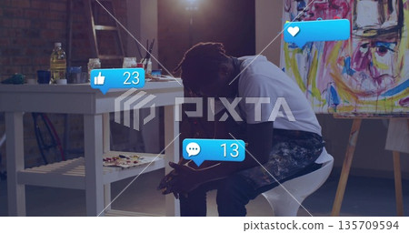 Leaning forward man holding smartphone on stool in studio with table, paint jars and floating icons 135709594