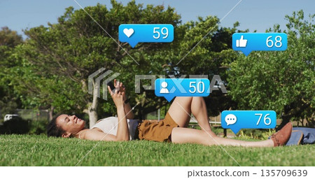 Lying woman wearing ribbed top and shorts holding smartphone in grassy park, with small blue bag 135709639