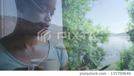 Standing woman wearing teal top holding white mug and smartphone by window viewing lake, copy space Standing woman wearing teal top holding white mug and smartphone by window viewing lake, copy space 135709646