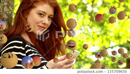 Redhead woman leaning against trunk holding smartphone gazing at camera in park, with emoji spheres Redhead woman leaning against trunk holding smartphone gazing at camera in park, with emoji spheres 135709650