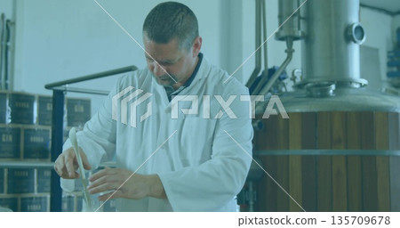 Stirring technician in white lab coat mixing clear liquid in flask at distillery, gin boxes, tank Stirring technician in white lab coat mixing clear liquid in flask at distillery, gin boxes, tank 135709678