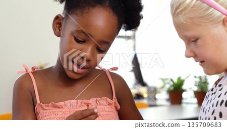Inspecting two friends holding small craft piece at bright play area table, with potted plants Inspecting two friends holding small craft piece at bright play area table, with potted plants 135709683