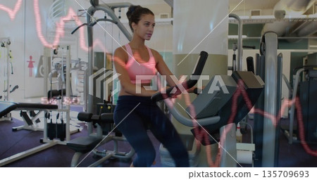 Pedaling woman wearing pink sportswear on exercise bike in gym, with heart-rate graph overlay 135709693