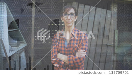 Standing mature woman crossing arms in front of barn wire mesh barrier, with metal fan housing 135709701
