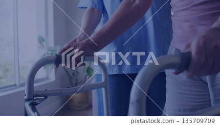 Supporting caregiver in scrubs assisting senior with walker in living area, with plant near window Supporting caregiver in scrubs assisting senior with walker in living area, with plant near window 135709709