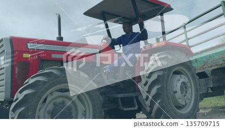 Driving operator wearing blue coveralls on dirt track, with red tractor and green metal trailer Driving operator wearing blue coveralls on dirt track, with red tractor and green metal trailer 135709715