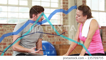 Holding clipboard instructor discussing workout with client in studio, cardio machine, towel Holding clipboard instructor discussing workout with client in studio, cardio machine, towel 135709735
