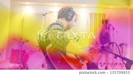 Recording musician strumming electric guitar in recording studio with drum kit left, vocalist right Recording musician strumming electric guitar in recording studio with drum kit left, vocalist right 135709833