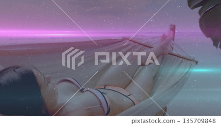 Relaxing adult woman wearing two-piece swimsuit on rope hammock at beach at dusk, with palm frond Relaxing adult woman wearing two-piece swimsuit on rope hammock at beach at dusk, with palm frond 135709848