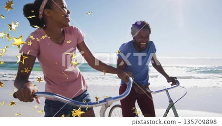 Walking couple wearing pink, blue T-shirts and shorts pushing bicycles along wet shoreline, waves Walking couple wearing pink, blue T-shirts and shorts pushing bicycles along wet shoreline, waves 135709898
