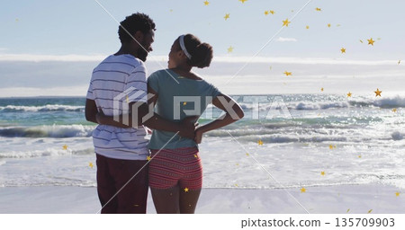 Embracing couple gazing at ocean waves on sandy shoreline, wearing striped shirt, patterned shorts Embracing couple gazing at ocean waves on sandy shoreline, wearing striped shirt, patterned shorts 135709903