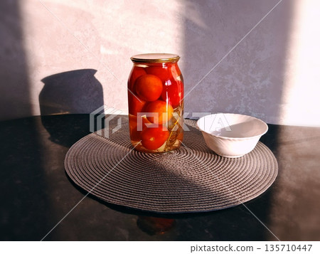 Jar of Pickled Tomatoes on Table Still Life. Glass jar filled with pickled cherry tomatoes in brine with herbs on dark table with bowl, warm lighting, home food preservation scene. 135710447