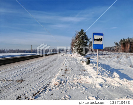 Snowy Rural Bus Stop Sign in Winter Landscape. Blue bus stop sign with van icon on snowy roadside in rural area, pine forest and empty winter road, seasonal travel scene. Snowy Rural Bus Stop Sign in Winter Landscape. Blue bus stop sign with van icon on snowy roadside in rural area, pine forest and empty winter road, seasonal travel scene. 135710488