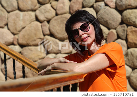 Elegant woman in orange dress standing against rustic stone wall, wearing sunglasses outdoors 135710781