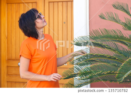 An adult woman in a short bright dress in the garden of her house near a palm tree An adult woman in a short bright dress in the garden of her house near a palm tree 135710782