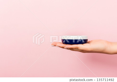 Female hand holding cream bottle of lotion isolated. Girl give jar cosmetic products on pink background 135711492
