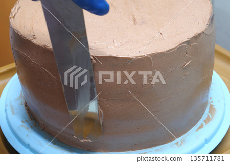 Confectioner woman is smearing cream on sides of cake using spatula, closeup view. culinary, baking and cooking food concept, home cooking Confectioner woman is smearing cream on sides of cake using spatula, closeup view. culinary, baking and cooking food concept, home cooking 135711781