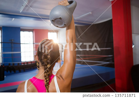 Female wearing pink top and leggings lifting gray kettlebell in boxing ring with large windows Female wearing pink top and leggings lifting gray kettlebell in boxing ring with large windows 135711871