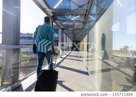 African American man walking along elevated glass corridor carrying suitcase and shoulder bag African American man walking along elevated glass corridor carrying suitcase and shoulder bag 135711884