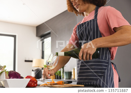 African American woman wearing apron pouring wine into glass at kitchen counter, copy space 135711887