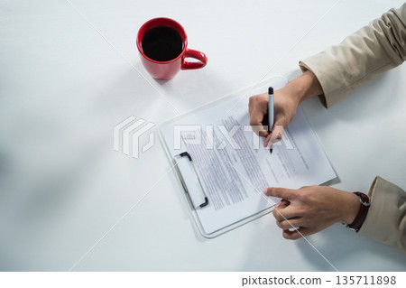 Asian woman signing printed document on clear clipboard at office desk with red mug, copy space 135711898