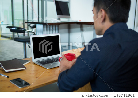 Mid adult Asian man holding red coffee mug and working on laptop in office, copy space Mid adult Asian man holding red coffee mug and working on laptop in office, copy space 135711901