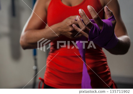Female athlete wearing red tank top and black shorts wrapping hands with purple wraps at gym Female athlete wearing red tank top and black shorts wrapping hands with purple wraps at gym 135711902