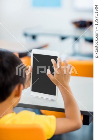 White tablet being held upright on white desk with blank screen in classroom, with orange chairs White tablet being held upright on white desk with blank screen in classroom, with orange chairs 135711904