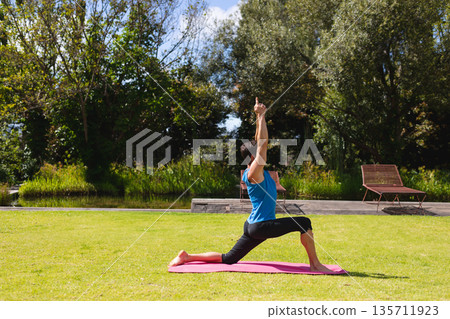 Man practicing lunge on pink mat stretching on grass by pond with wooden deck lounge chairs Man practicing lunge on pink mat stretching on grass by pond with wooden deck lounge chairs 135711923