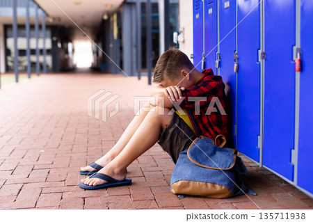 School-aged boy sitting on school corridor leaning against blue lockers with backpack, copy space 135711938