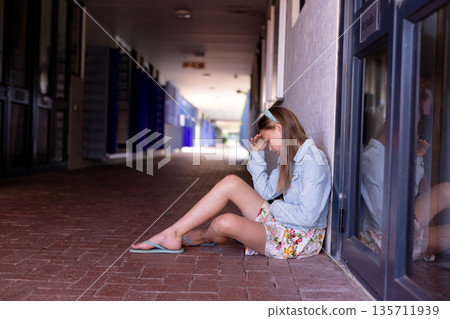 Girl sitting on brick arcade beside glass door sign, windows wearing denim jacket, copy space 135711939