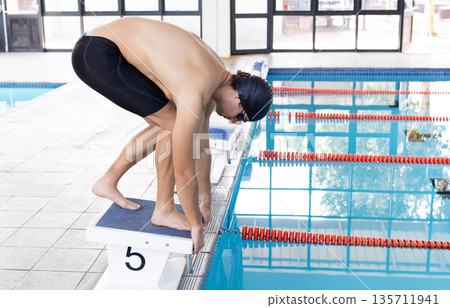 Male athlete crouching on starting block at lap pool wearing swim cap and goggles, copy space 135711941