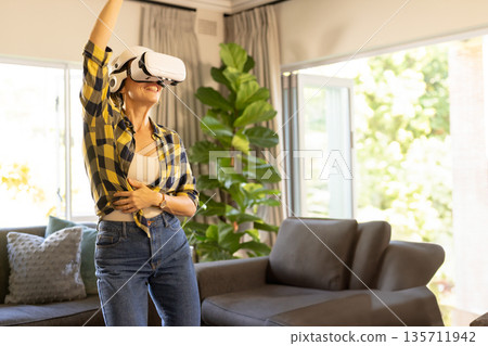 Woman wearing VR headset raising arm while interacting in living room with potted fiddle leaf fig 135711942