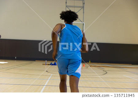 African American male basketball player running toward hoop at gym near orange cones, water bottle African American male basketball player running toward hoop at gym near orange cones, water bottle 135711945