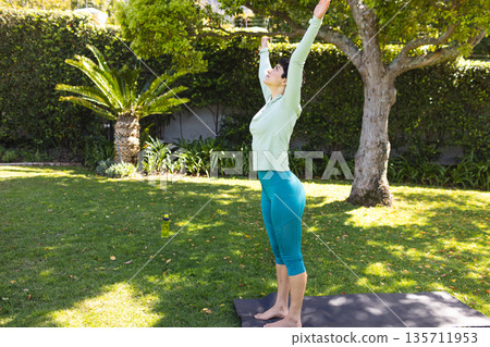 Female practicing yoga on lawn in private backyard with yoga mat and water bottle, copy space 135711953